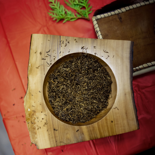 A light-colored wooden bowl with a round center and square outer edges filled with Semaa (Tobacco for Anishinaabe ceremonial use) set on a reddish-orange background with a leaf from a White Cedar Tree and part of a rectangular brown birch bark item with a white stitched border visible next to the bowl.